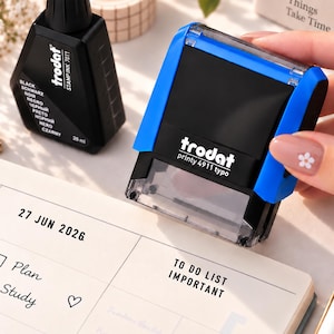 May include: A blue and black Trodat Printy 4911 typo self-inking stamp positioned above a planner page with the text 27 JUN 2026 and TO DO LIST IMPORTANT. A black bottle of Trodat stamp ink 7011 is visible in the background.