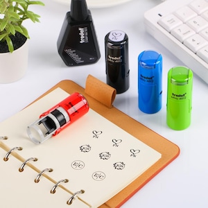 May include: A collection of Trodat self-inking stamps in various colors, including red, black, blue, and green, alongside a bottle of black stamp ink. The stamps have the text "trodat 4612" and are shown with stamped impressions on a notepad.