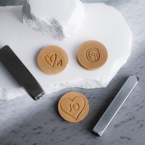 May include: A collection of round, tan-colored objects with embossed designs, including hearts and initials, alongside two silver metal tools. The objects are arranged on a white, textured surface with a gray marble background.