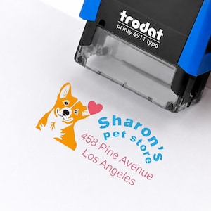 May include: A black and blue Trodat Printy 4911 typo self-inking stamp. The stamp impression features an orange and white dog illustration, a pink heart, and the blue text Sharon's pet store above the red text 458 Pine Avenue Los Angeles.