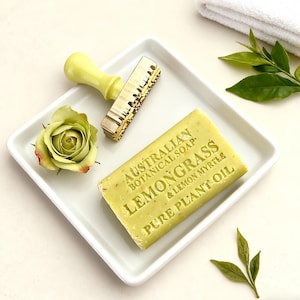 May include: A square white dish holds a light green bar of soap, a yellow stamp, and a green rose. The soap is labeled "Australian Botanical Soap Lemongrass & Lemon Myrtle Pure Plant Oil". A white towel and green leaves are in the background.