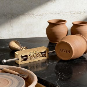 May include: A brass stamp with the name "KEVIN" and the numbers "8-6-50" is shown next to a terracotta pot with the name "KEVIN" stamped on the bottom. Two other terracotta pots are in the background. A brass handle is also visible.