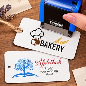 May include: A blue and black Trodat Printy 4911 typo self-inking stamp pressing onto a white rectangular tag with a bakery logo featuring a chef hat, bread, and wheat. Below, a second white tag displays a blue tree icon and the text Abdelhak Enjoy your reading time!