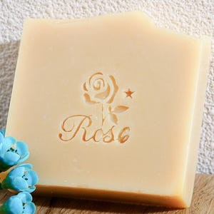 Peut inclure: Un pain de savon carré beige clair avec un motif de rose et le mot "Rose" gravé sur la surface. Une petite étoile est à côté de la rose. Le savon est de couleur naturelle et a une texture lisse. Le savon est un produit de beauté.
