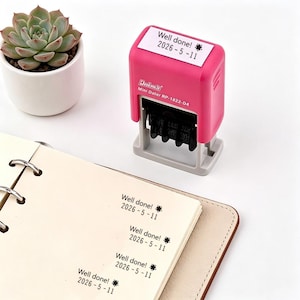 May include: A pink and grey self-inking stamp, marked "Well done! 2026-5-11", sits beside a notebook with the same stamped text. A small succulent in a white pot is in the background.