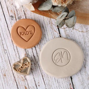 May include: Three custom stamps on a wooden surface. One is a heart-shaped stamp with the text "KAO'S" inside. Another is a round stamp with the initials "EM" inside a circle. The third is a heart-shaped metal stamp with the text "SOAY".