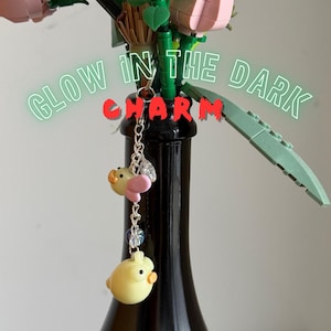 Peut inclure: Une breloque en chaîne métallique argentée avec deux petites figurines en forme de canard jaune, bec orange et détail d'aile rose, agrémentée d'une perle transparente. Le texte GLOW IN THE DARK CHARM est superposé en vert et rouge.