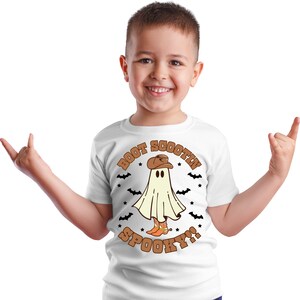 以下が含まれることがあります： カウボーイハットとブーツを履いた幽霊が描かれた白いTシャツ。Tシャツには「Boot Scootin' Spooky!」の文字がプリントされています。