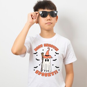 May include: White t-shirt with an orange graphic of a ghost wearing a witch hat and holding a broom. The text "BOOT SKOOTIN SPOOKY!" is printed on the shirt.