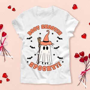Peut inclure: T-shirt blanc avec un graphique d'un fantôme portant un chapeau de sorcière et tenant un balai. Le texte "Boot Scootin' Spooky!" est imprimé sur le t-shirt.