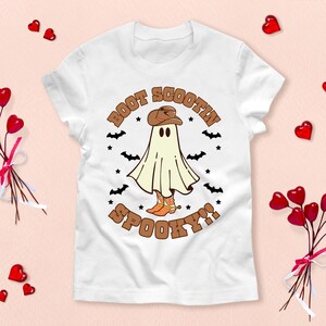 Peut inclure: T-shirt blanc avec un fantôme portant un chapeau de cowboy et des bottes. Le texte "Boot Scootin' Spooky!" est imprimé sur le t-shirt.
