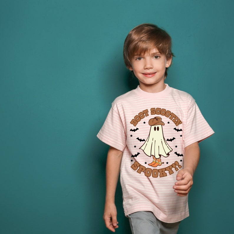 以下が含まれることがあります： カウボーイハットとブーツを履いた幽霊が描かれたピンクのストライプのTシャツ。Tシャツには「Boot Scootin' Spooky!」の文字が書かれています。