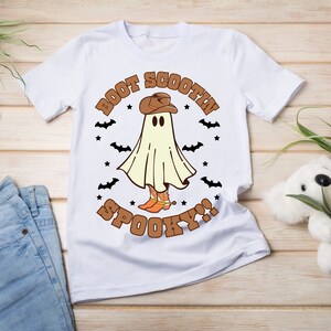 以下が含まれることがあります： カウボーイハットとブーツを履いた幽霊が描かれた白いTシャツ。Tシャツには「Boot Scootin' Spooky!」の文字がプリントされています。