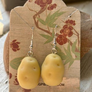 Puede incluir: Un par de pendientes colgantes con dijes realistas en forma de patata de color tostado claro, suspendidos de ganchos de metal plateado. Los pendientes se presentan en una tarjeta decorativa con un estampado floral y de bambú.