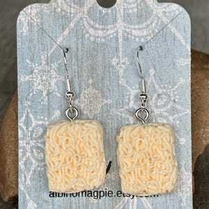 May include: Novelty earrings shaped like miniature ramen noodle blocks. The cream-coloured earrings have detailed noodle strands and hang from silver-coloured fish hook ear wires. They are displayed on a light blue tag with a snowflake pattern.