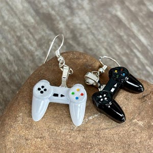 Peut inclure: Paire de boucles d'oreilles représentant des manettes de jeu miniatures. L'une est blanche avec des boutons colorés, l'autre noire. Les deux ont des crochets d'oreilles argentés. Elles ressemblent à des manettes de jeux vidéo classiques.