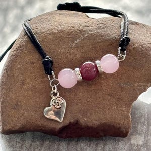 Puede incluir: Una pulsera hecha a mano con cuentas de piedras preciosas rosas y burdeos, espaciadores plateados y un dije de corazón plateado con un diseño de rosa. La pulsera está ensartada en un cordón negro y se muestra sobre una superficie marrón texturizada.