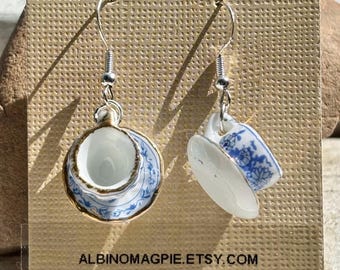 May include: A pair of dangling earrings featuring miniature teacups and saucers. The white porcelain pieces are adorned with blue floral patterns and gold trim. The earrings are suspended from silver-colored hooks. The text "ALBINOMAGPIE.ETSY.COM" is visible at the bottom.