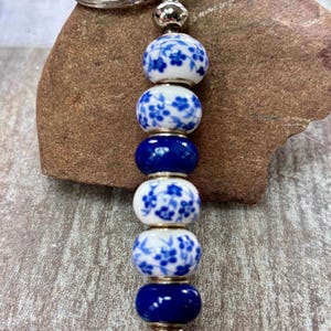 Op de afbeelding: Een sleutelhanger met een zilverkleurige metalen ring en een verticale rij van zes kralen. De kralen wisselen af tussen effen donkerblauwe en witte keramische bollen met een blauw bloemmotief, gescheiden door kleine zilverkleurige tussenstukjes.