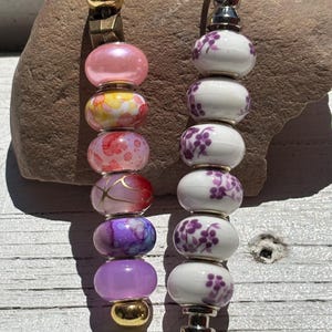 May include: Two keychains featuring vertical arrangements of beads. The left keychain has multicolored glass beads in pink, yellow, and purple hues with gold-tone hardware. The right keychain features white ceramic beads with purple floral patterns.