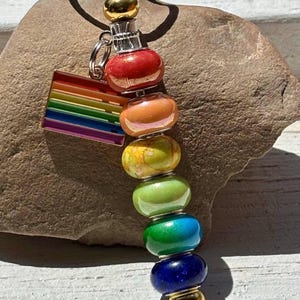 Op de afbeelding: Een kleurrijke sleutelhanger met een regenboogvlagbedel en een reeks levendige, kralen bollen in rood, oranje, geel, groen, blauw en paars. De sleutelhanger heeft een goudkleurige bovenkant en een zilverkleurige ring.
