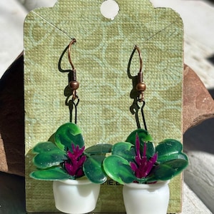 May include: A pair of dangle earrings featuring miniature white pots containing green succulent plants with vibrant purple centers. The earrings are attached to copper-toned metal hooks and are displayed on a textured light green cardstock backing.