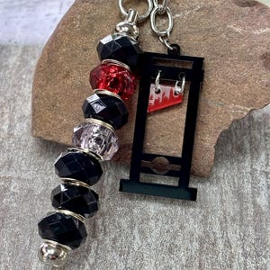 May include: A keyring featuring a silver-toned metal ring and chain attached to a vertical row of faceted beads in black, red, and clear colours, alongside a black guillotine-shaped charm with a small red triangular blade accent.
