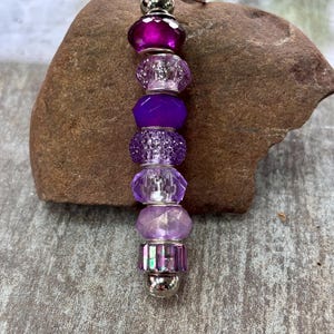 Op de afbeelding: Een sleutelhanger met een verticale rangschikking van zeven kralen in verschillende tinten paars en magenta. De kralen hebben gefacetteerde, getextureerde en gladde stijlen, gescheiden door kleine zilverkleurige metalen tussenstukjes en een ring.