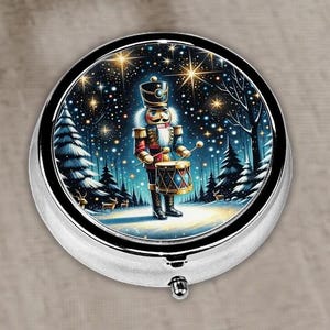 May include: A circular, silver pill box with a winter scene depicting a nutcracker playing a drum in a snowy forest. The design includes stars, fir trees, and deer, evoking a festive Christmas theme.