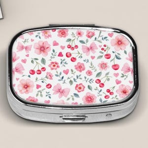 Puede incluir: Una caja de pastillas de plata con fondo blanco y un patrón floral rosa con cerezas y corazones.