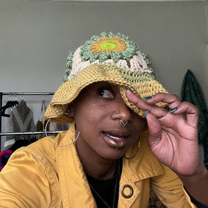 May include: A handmade crochet bucket hat with a sunflower design. The hat features a yellow brim, a cream-colored base, and a green and yellow sunflower on top. The hat is a unique accessory.