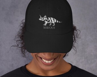 May include: Black baseball cap featuring a white and gray triceratops illustration. The word "SERIOUS" is printed below the dinosaur graphic. The cap has a curved brim and a structured fit.