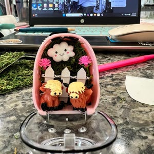 May include: A pink, decorative soap dish with a clear, round soap holder. Inside the dish is a miniature scene with two brown and yellow Highland cows, a white picket fence, and a smiling cloud. Pink flowers and green moss complete the scene.