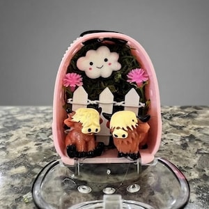 Może przedstawiać: Różowy pojemnik w kształcie jajka zawierający miniaturową scenkę z dwiema brązowymi figurkami krów rasy Highland, białym płotkiem, dwoma różowymi kwiatami i białą uśmiechniętą chmurką na tle ciemnozielonego mchu.