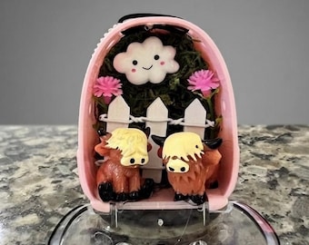 May include: A pink, egg-shaped container holding a miniature scene with two brown Highland cow figurines, a white picket fence, two pink flowers, and a white smiling cloud figure set against a dark green mossy background.