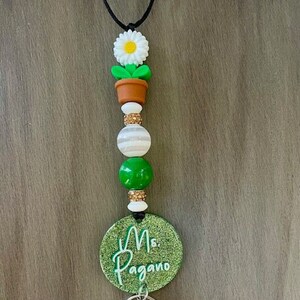 May include: A green and white beaded lanyard with a flower charm, a round green tag with the text "Ms. Pagano", and a key chain with a tassel.