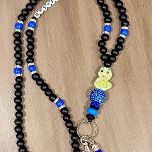 Peut inclure: Un cordon porte-badge composé de perles noires mates, de perles bleues vives et d'entretoises argentées. Il comprend des perles lettres blanches formant MRS FERRARO avec un cœur bleu, une breloque serpent vert clair, une perle strass et un pompon bleu.