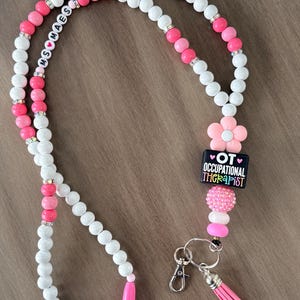 May include: A beaded lanyard featuring white and pink round beads, silver-toned spacers, and a pink flower charm. It includes a black square bead with the text "OT OCCUPATIONAL THERAPIST" and a white bead segment reading "MS MAES" with a heart icon, finished with a pink tassel.