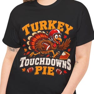 Pode incluir: Uma t-shirt preta com um gráfico de um peru a usar um capacete de futebol americano e a correr com uma bola. O texto diz TURKEY TOUCHDOWNS PIE, acompanhado por ilustrações de bolas, folhas de outono e uma fatia de tarte na parte inferior.
