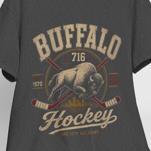 May include: Dark gray t-shirt with a vintage-style Buffalo Hockey design. The design includes the word "Buffalo", the number "716", a buffalo illustration, crossed hockey sticks, and the words "Hockey" and "One City, All Heart."