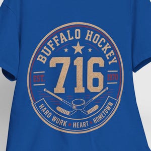 Op de afbeelding: Koningsblauw T-shirt met een cirkelvormige, bruine afbeelding met de tekst "BUFFALO HOCKEY" en "716". Het ontwerp bevat gekruiste hockeysticks en de woorden "HARD WORK HEART HOMETOWN". Een witte baseballpet ligt op een houten stoel.