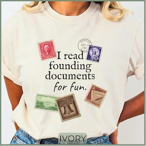 May include: An ivory t-shirt featuring the black text I read founding documents for fun. The design includes five vintage-style postage stamps in red, purple, green, brown, and tan, along with a circular postmark dated September 17, 1787.