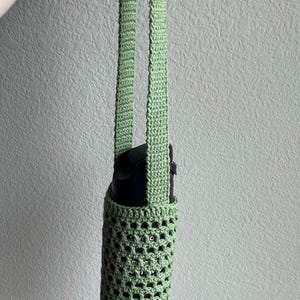 May include: A green crocheted water bottle holder with a long strap. The holder is made of a delicate, open-weave pattern.