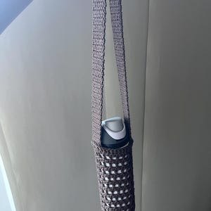 Op de afbeelding: Een bruine gehaakte waterfleshouder met een lange band. De houder is gemaakt met een eenvoudig steekpatroon en heeft een witte waterfles erin.