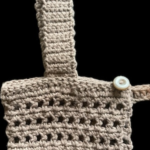 Op de afbeelding: Een bruine gehaakte tas met een enkele knoopsluiting. De tas heeft een geweven patroon en een lange band.