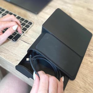 May include: A black desk organizer with a drawer holding a charging cable. The organizer is on a wooden desk, with a laptop and keyboard in the background. The drawer is open, revealing the cable.