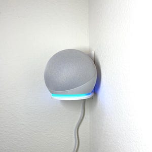 Peut inclure: Un haut-parleur intelligent sphérique gris monté sur un support mural blanc. Le haut-parleur a une texture semblable à du tissu et se trouve au-dessus d'un anneau lumineux bleu. Un cordon d'alimentation gris s'étend le long du mur.