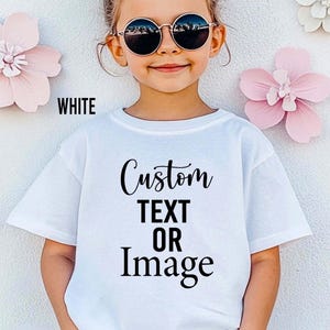 May include: A white t-shirt with the words "Custom TEXT OR Image" in black script. The word "WHITE" is printed in black on the upper left. The shirt is being worn by a young person wearing round sunglasses.