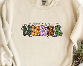 May include: Sand-colored sweatshirt with the word "NURSE" in colorful, patterned letters. The design includes bats, spiderwebs, and a nurse's hat, suggesting a Halloween theme. The sweatshirt is laid flat with jeans and white sneakers.