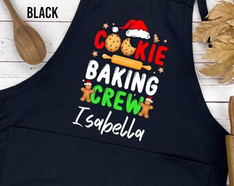 May include: Black apron with a large front pocket. The apron features the text "COOKIE BAKING CREW Isabella" in a festive design with cookies, a rolling pin, and gingerbread men. A wooden spoon and rolling pin are on the sides.